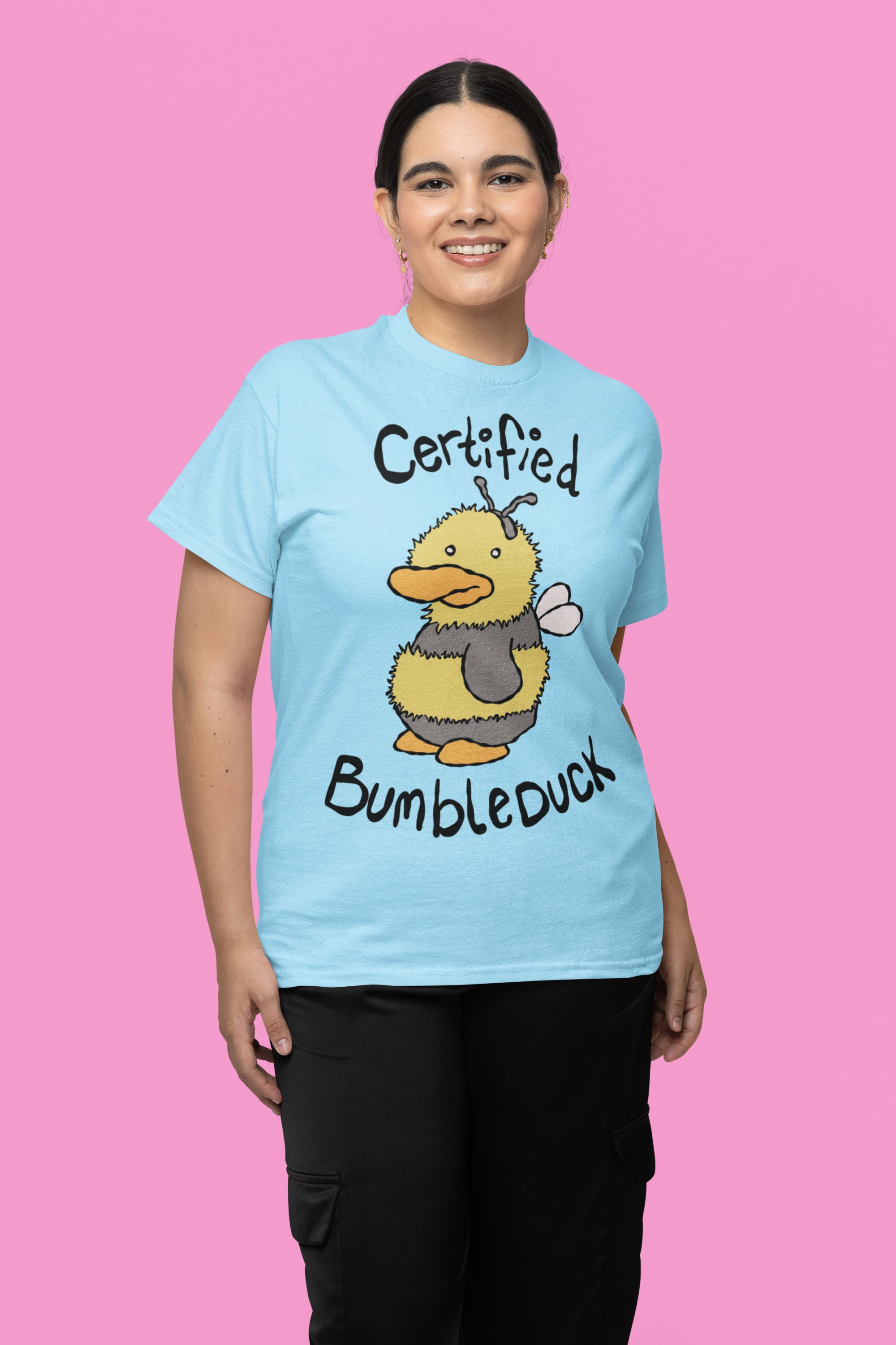 Woman wearing a light blue t-shirt with a cartoon bee and text CERTIFIED BUMBLEDUCK, standing in front of pink background