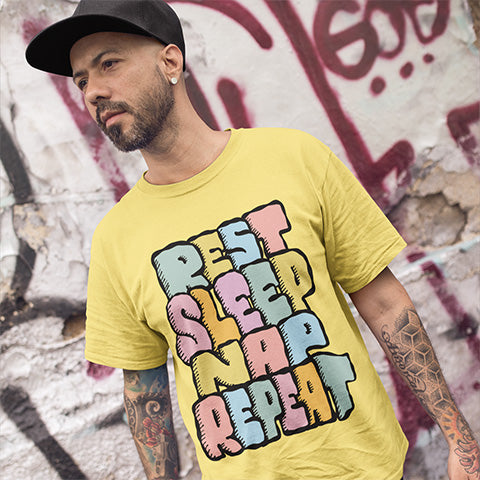 Man wearing a yellow t-shirt with colourful text REST SLEEP NAP REPEAT design in front of a graffiti-covered wall.